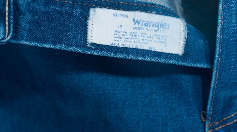 May include: Close-up of blue denim jeans with a white label. The label reads "Wrangler" and includes washing instructions and size 12. Made in the USA.
