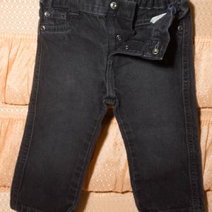 May include: Dark gray denim pants with a button and snap closure. The pants have a small pocket on the front and a comfortable elastic waistband. The denim is slightly faded, giving it a worn look.