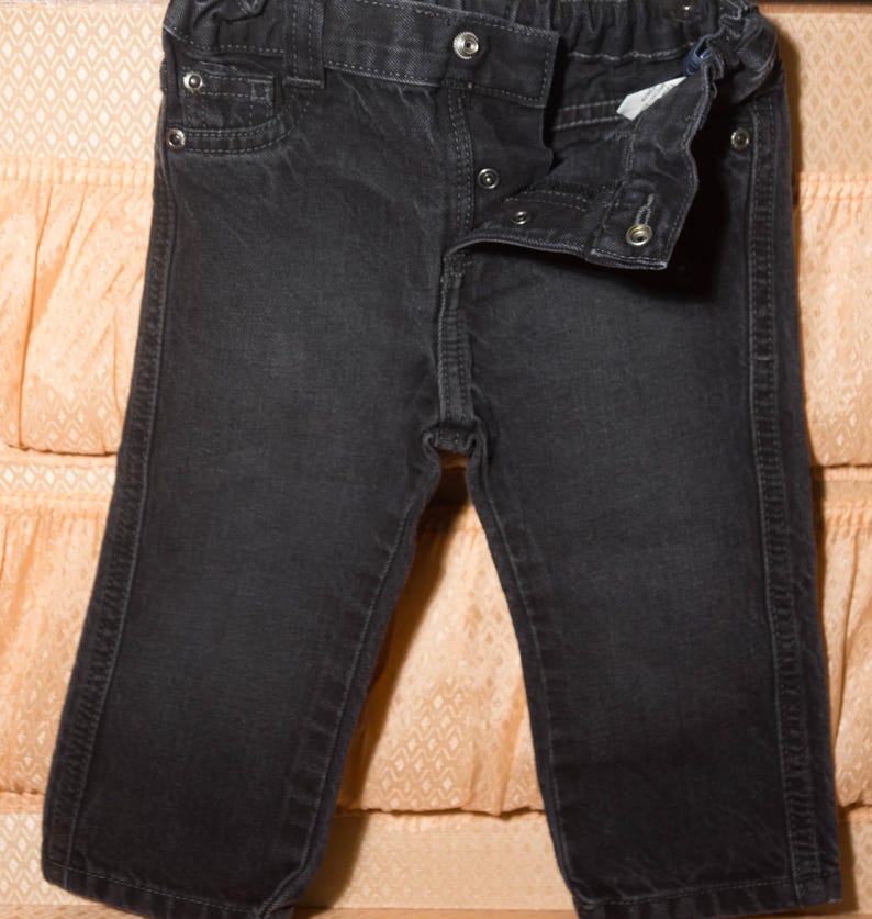 May include: Dark gray denim pants with a button and snap closure. The pants have a small pocket on the front and a comfortable elastic waistband. The denim is slightly faded, giving it a worn look.