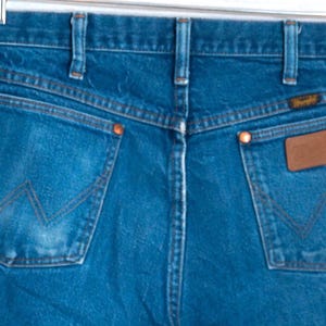 Puede incluir: Vista trasera de unos vaqueros azules. Los vaqueros tienen dos bolsillos traseros con un dise&ntilde;o de costura en forma de "W". Un parche de cuero marr&oacute;n est&aacute; en el bolsillo derecho y la etiqueta de la marca Wrangler es visible.