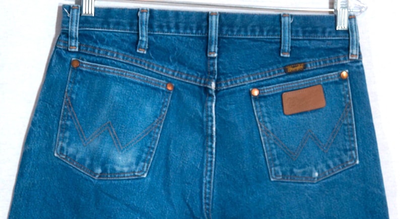 Puede incluir: Vista trasera de unos vaqueros azules. Los vaqueros tienen dos bolsillos traseros con un dise&ntilde;o de costura en forma de "W". Un parche de cuero marr&oacute;n est&aacute; en el bolsillo derecho y la etiqueta de la marca Wrangler es visible.