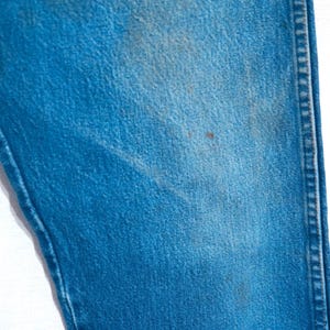 Puede incluir: Primer plano de una secci&oacute;n de vaqueros azules. La tela muestra desgaste con manchas y decoloraci&oacute;n visibles. La costura a lo largo de la costura es visible, y la textura general es &aacute;spera.
