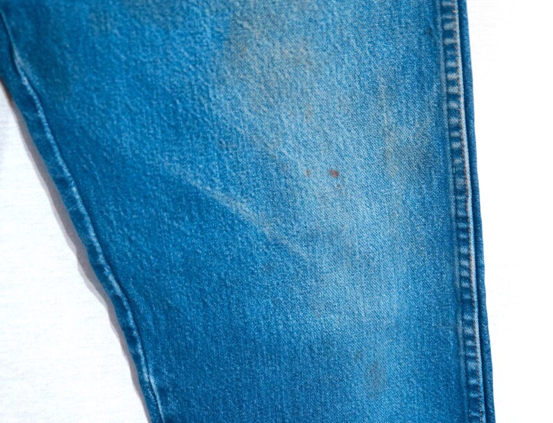 Puede incluir: Primer plano de una secci&oacute;n de vaqueros azules. La tela muestra desgaste con manchas y decoloraci&oacute;n visibles. La costura a lo largo de la costura es visible, y la textura general es &aacute;spera.