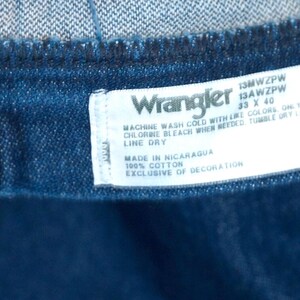 May include: Close-up of a dark blue denim garment with a white label. The label reads "Wrangler" and includes washing instructions, size 33 x 40, and "Made in Nicaragua." The garment is 100% cotton.