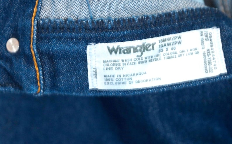May include: Close-up of a dark blue denim garment with a white label. The label reads "Wrangler" and includes washing instructions, size 33 x 40, and "Made in Nicaragua." The garment is 100% cotton.
