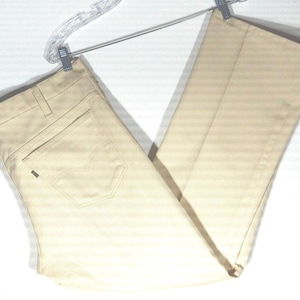 Puede incluir: Un par de vaqueros Levi's de color beige claro colgados de una percha transparente. Los vaqueros tienen un diseño clásico de cinco bolsillos, con un bolsillo trasero visible. El logotipo de Levi's está en el bolsillo trasero. Los pantalones son de tela resistente.