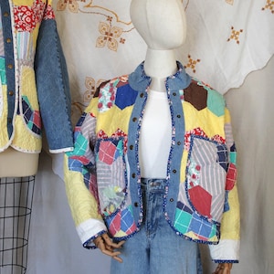 Puede incluir: Una chaqueta de patchwork confeccionada con tela acolchada que presenta un patrón hexagonal en amarillo, rojo, azul y blanco, acentuada con mangas y ribetes de mezclilla azul. La chaqueta tiene cuello alzado, cierre frontal de botones y dos bolsillos.