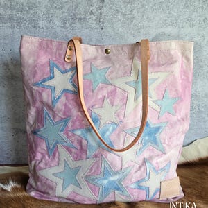 Puede incluir: Un bolso tote con base de tela rosa teñida con efecto tie-dye y múltiples estrellas aplicadas en tonos azul claro y crema. El bolso incluye dos correas de hombro de cuero color canela sujetas con remaches de latón y una etiqueta de cuero con INTIKA.