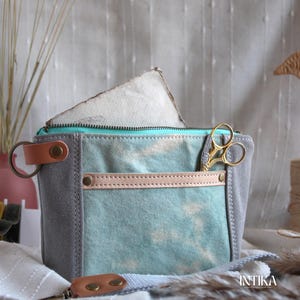 May include: A gray and teal canvas pouch with a leather accent and a teal zipper. Gold scissors are tucked into the side pocket. The pouch is open, revealing a white object inside. The word "INTIKA" is visible.