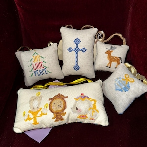 May include: Six small, white, fabric ornaments with embroidered designs. The ornaments feature a cross, a deer, a gift box, a Christmas tree with the words "Joy, Love, Peace", and a scene from Beauty and the Beast with Lumiere, Cogsworth, and Mrs. Potts.
