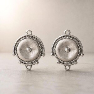 Puede incluir: Dos conectores de joyería circulares de metal plateado con un punto central en relieve, un borde de cuentas y pequeñas anillas en la parte superior e inferior para su fijación. El metal tiene un acabado mate ligeramente desgastado.