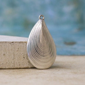 May include: A silver-toned metal pendant shaped like a teardrop or mussel shell, featuring a series of concentric, etched lines that follow the curved outline of the piece. A small, circular jump ring is attached to the top for hanging purposes.
