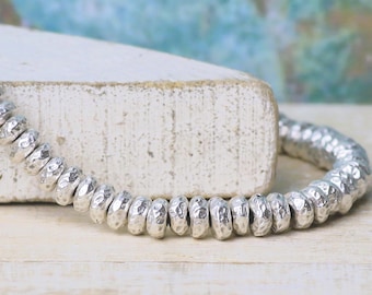 May include: A strand of small, round, silver-colored beads with a textured, hammered finish. The beads are strung together in a continuous line, showcasing a metallic sheen and a consistent, slightly irregular surface pattern on each individual bead.