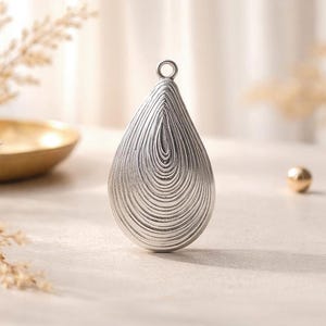 May include: A silver pendant in a teardrop shape, featuring a textured surface with concentric, curved lines. It has a small loop at the top for hanging. A gold bowl and a small gold sphere are in the background.