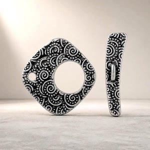 Könnte beinhalten: Ein Knebelverschluss-Set aus silberfarbenem Metall, bestehend aus einem quadratischen Ring mit kreisförmigem Ausschnitt und einem passenden gebogenen Stab. Beide Teile weisen ein komplexes, erhabenes Muster aus Wirbeln und kleinen Punkten auf.