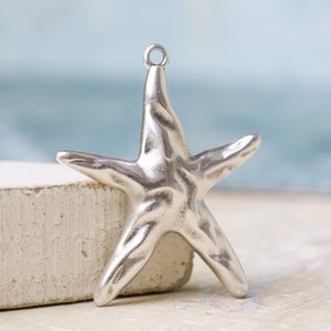 May include: A silver-toned metal starfish pendant featuring a textured, uneven surface and a small circular jump ring at the top for attachment. The starfish has five distinct arms and a reflective, metallic finish with visible light highlights.