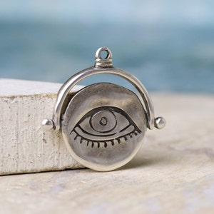 Könnte beinhalten: Ein silberner Anhänger mit einem drehenden Augenmotiv. Das Auge ist mit einer Pupille und Wimpern detailliert. Der Anhänger ist an einem silbernen Ring befestigt.
