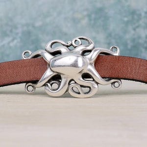 Puede incluir: Una pulsera de cuero marrón con un dije central de metal plateado en forma de pulpo. El pulpo tiene una cabeza lisa y redondeada con ocho tentáculos curvos que se extienden horizontalmente sobre la banda de cuero, asegurada por pequeñas anillas.