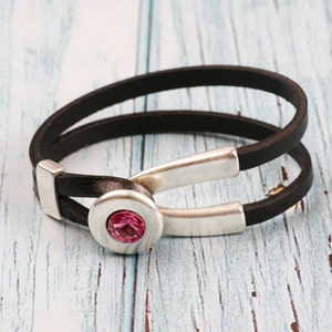 Puede incluir: Un brazalete de cuero negro con cierre de plata y un acento de cristal rosa.