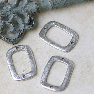 Pu&ograve; includere: Tre ciondoli rettangolari in metallo argentato con una superficie testurizzata e due fori ciascuno. I ciondoli misurano circa 2,5 cm di lunghezza e 2 cm di larghezza.