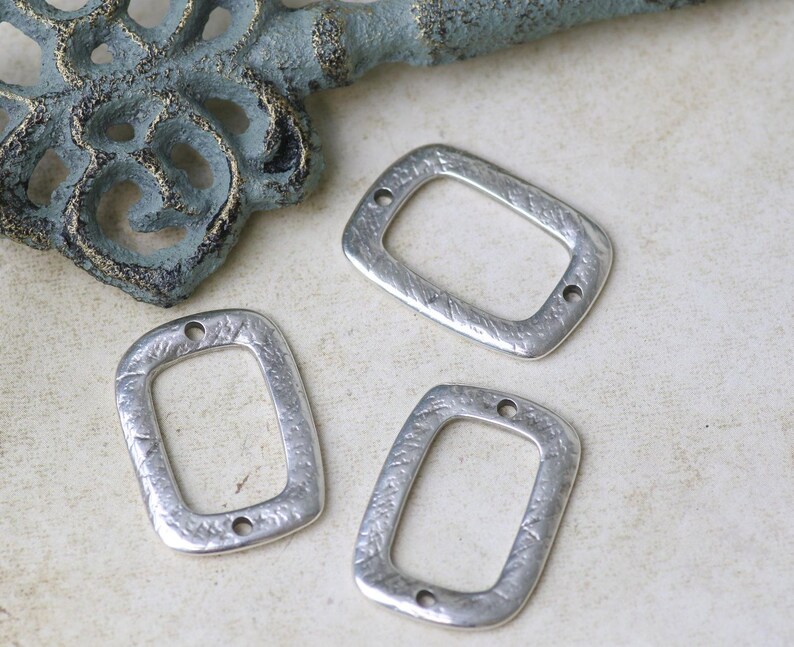 Pu&ograve; includere: Tre ciondoli rettangolari in metallo argentato con una superficie testurizzata e due fori ciascuno. I ciondoli misurano circa 2,5 cm di lunghezza e 2 cm di larghezza.