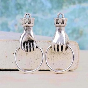 Könnte beinhalten: Zwei silberfarbene Metallanhänger, die jeweils eine Hand darstellen, die einen kreisförmigen Ring hält. Der Handgelenksbereich jeder Hand ist mit einem dekorativen Band mit kleinen blattartigen Gravuren versehen. Die Anhänger haben ein Metallfinish.