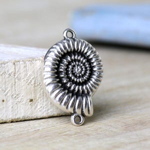 Könnte beinhalten: Ein silberfarbener Metall-Verbindungsanhänger in Form eines Ammoniten-Fossils mit spiralförmigem Muschelmuster. Der Anhänger verfügt über eine kreisförmige Öse an der Ober- und Unterseite zur Befestigung sowie eine polierte Oberfläche.