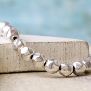 Pode incluir: Um bracelete de prata com contas facetadas. As contas são redondas e têm uma textura martelada. O bracelete está sobre uma superfície branca.