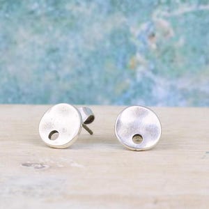 May include: Two silver-colored stud earrings. Each earring has a flat, round face with a small, centered hole. The earrings are set against a blurred, light blue and green background.