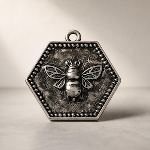 Può includere: Un ciondolo esagonale in metallo color argento con un motivo a ape in rilievo al centro. Il bordo dell'esagono è decorato con una trama a perline. La superficie presenta una finitura scura e anticata, con un piccolo occhiello circolare in alto.
