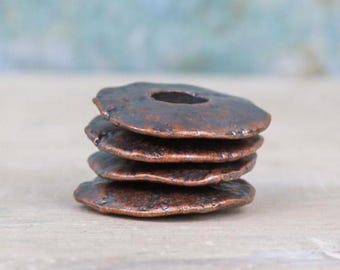 May include: A stack of four circular, flat, copper-colored metal beads with a dark, weathered, and textured finish. Each bead features a central hole and irregular, organic edges, creating a rustic appearance with varying tones of brown and bronze.