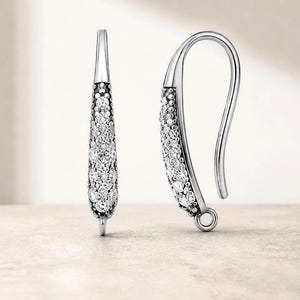 May include: A pair of silver-toned earring hooks featuring a curved, elongated teardrop shape. The front surface of each earring is encrusted with numerous small, clear, sparkling stones set against a dark, textured background.