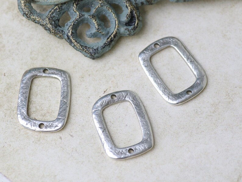 Pu&ograve; includere: Tre ciondoli rettangolari in metallo argentato con superficie testurizzata e due fori nella parte superiore.