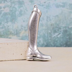 May include: A silver-toned metal charm shaped like a tall equestrian riding boot. The charm features a smooth, polished finish with a small loop at the top for attachment. The design includes a defined heel, a rounded toe, and a structured shaft.