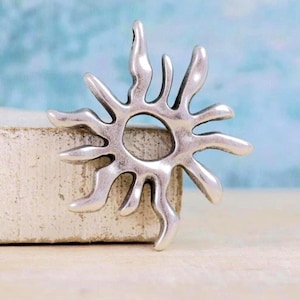 May include: A silver-coloured metal pendant shaped like a stylised sun with a circular centre and multiple radiating, wavy, tapered points. The surface has a smooth, polished finish with subtle darkened recessed areas to define the sunburst shape.