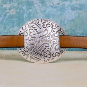 Könnte beinhalten: Ein brauner Ledergürtel mit einer großen, kreisförmigen Schnalle aus silberfarbenem Metall. Die Schnalle weist ein detailliertes Reliefmuster aus geschwungenen, blattartigen Formen auf, das einen Kontrast zum glatten, schlichten Lederriemen bildet.
