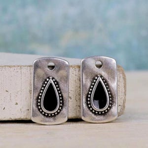 May include: Two rectangular silver-toned metal pendants featuring a central black teardrop-shaped inlay surrounded by a beaded border. Each pendant has a circular hole at the top for attachment, with a weathered, matte finish on the metal surface.