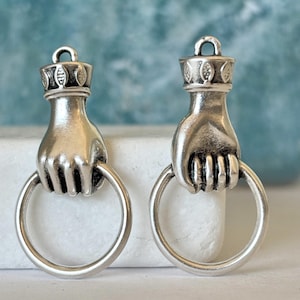 Könnte beinhalten: Zwei silberfarbene Metallanhänger, die jeweils eine Hand darstellen, die einen kreisförmigen Ring hält. Der Handgelenksbereich ist mit einem dekorativen Band mit geprägtem Blattmuster und einer kleinen Öse zur Befestigung versehen.