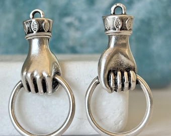 Könnte beinhalten: Zwei silberfarbene Metallanhänger, die jeweils eine Hand darstellen, die einen kreisförmigen Ring hält. Der Handgelenksbereich ist mit einem dekorativen Band mit geprägtem Blattmuster und einer kleinen Öse zur Befestigung versehen.