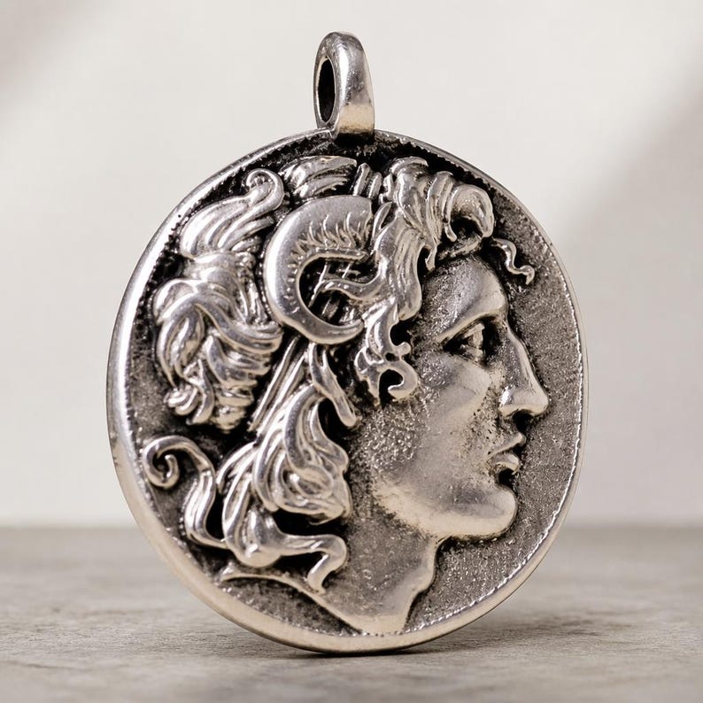 Op de afbeelding: Een ronde zilverkleurige hanger met een reli&euml;f van een portret in profiel van een figuur met krullend haar en een hoornachtig detail op het hoofd. Het metaal heeft een geoxideerde afwerking met donkere verdiepte delen en een gepolijst oogje.