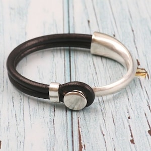Puede incluir: Una pulsera de cuero negro con cierre y detalles metálicos plateados. La pulsera presenta una banda de cuero de doble hebra y un diseño circular. Los componentes metálicos tienen un acabado pulido, creando un contraste con el cuero oscuro.