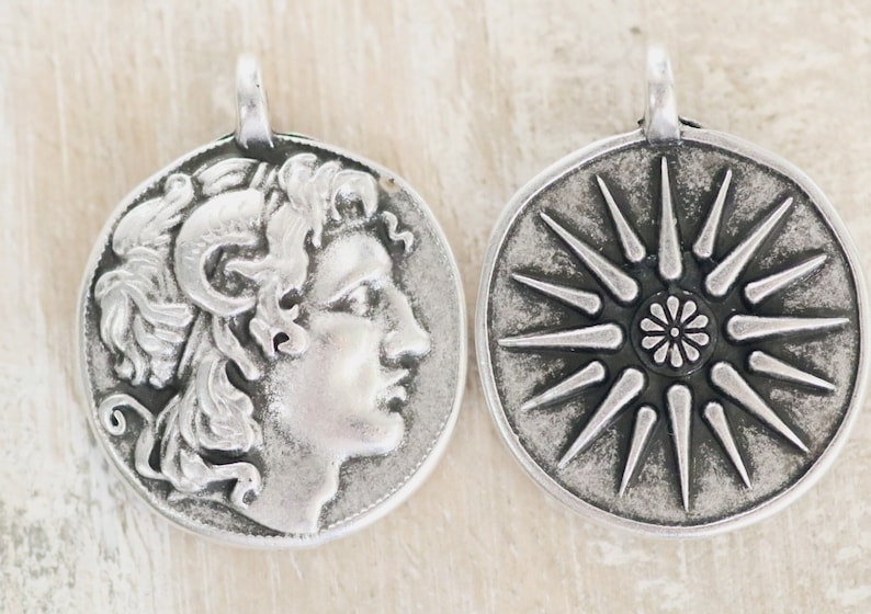 Op de afbeelding: Twee ronde hangers van zilverkleurig metaal met reli&euml;fontwerpen. De ene hanger toont het profiel van een figuur met krullend haar en ramshoorns, terwijl de andere een zestienpuntige ster met een centraal bloemmotief laat zien.