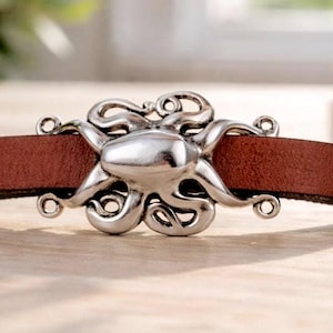 Könnte beinhalten: Ein braunes Lederband mit einer zentralen, silberfarbenen Metallschnalle in Form eines Oktopus. Die Schnalle hat eine polierte Oberfläche mit detaillierten Tentakeln, die sich horizontal über das Band erstrecken und einen Verschluss bilden.