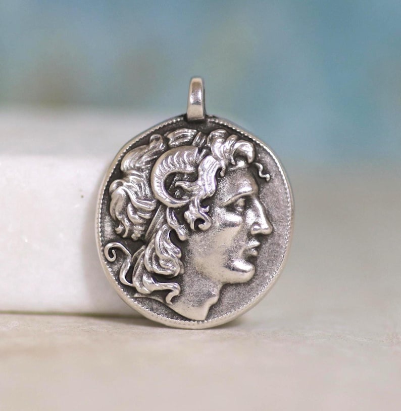 May include: A circular silver-toned pendant featuring a relief profile of a figure with curly hair and ram horns. The design is enclosed within a beaded border and includes a small integrated bail at the top for attachment to a chain or cord.