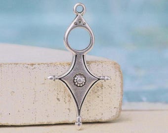 Op de afbeelding: Een zilverkleurige metalen hanger met een symmetrische, langwerpige kruisvorm, voorzien van een cirkelvormige lus aan de bovenkant, een centraal bloemachtig reliëfdetail en een klein kraalachtig accent aan de onderpunt.
