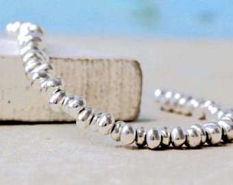 Puede incluir: Una pulsera de tono plateado compuesta por numerosas cuentas metálicas pequeñas y redondeadas ensartadas en una línea continua. Las cuentas tienen una superficie pulida y reflectante con formas orgánicas irregulares y texturizadas.