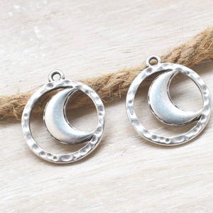 Op de afbeelding: Twee zilverkleurige metalen bedels, elk met een halve maanvorm gecentreerd in een cirkelvormig frame. Het cirkelvormige frame heeft een gehamerde, getextureerde afwerking en elke bedel bevat een klein lusje aan de bovenkant voor bevestiging.