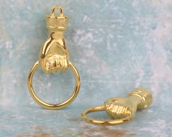 May include: Two gold-toned metal charms, each shaped like a hand holding a circular ring. One charm stands upright with a small loop at the top for attachment, while the other lies horizontally on its side, showcasing the metallic finish and form.