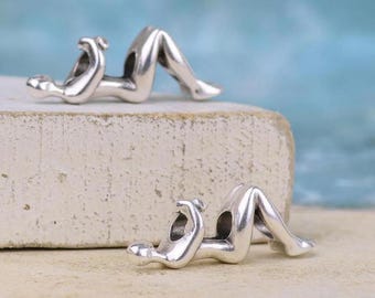 Könnte beinhalten: Zwei silberfarbene Metallanhänger, die jeweils eine stilisierte menschliche Figur in liegender Position mit angewinkelten Knien darstellen. Die Figuren haben glatte, polierte Oberflächen und eine hohle Öffnung im Rumpf zum Auffädeln.