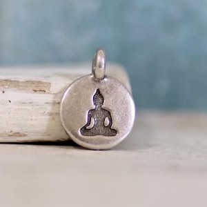 Peut inclure: Un pendentif circulaire en métal argenté présentant une silhouette en creux de couleur sombre représentant un Bouddha assis en position de méditation. Le pendentif possède une surface lisse légèrement patinée et une bélière intégrée.